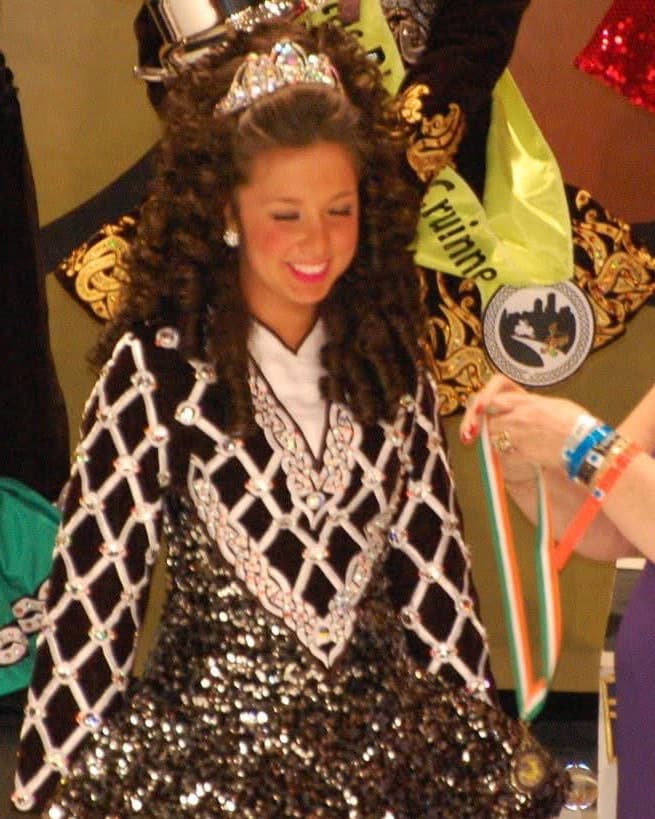 Liandra smiling on stage in a tiara and a black and white diamond-patterned Irish dance costume, being presented with a medal on a green, white and orange ribbon