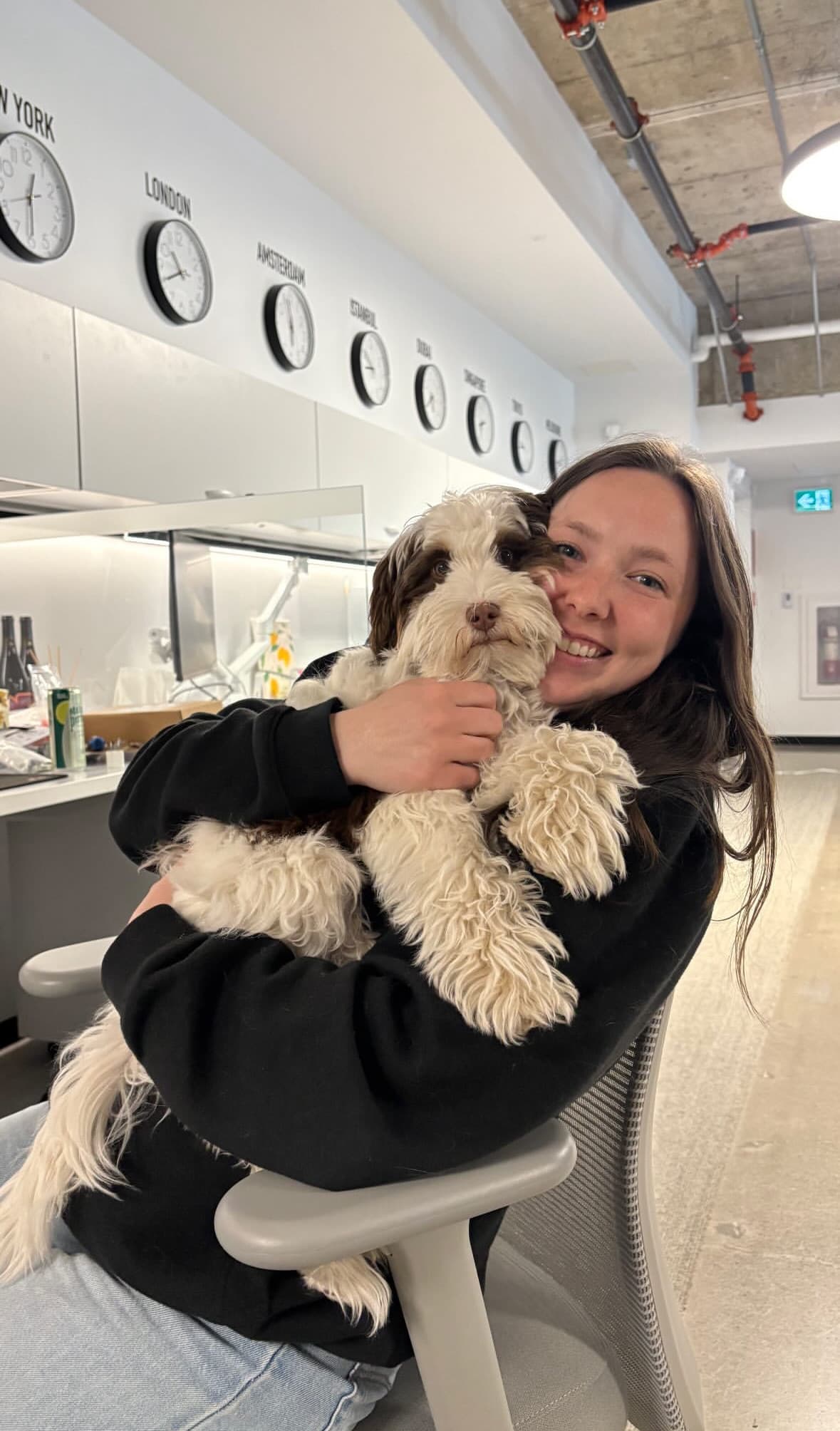 Liandra hugging a fluffy brown-and-white puppy in an office, with a row of world clocks on the wall behind her