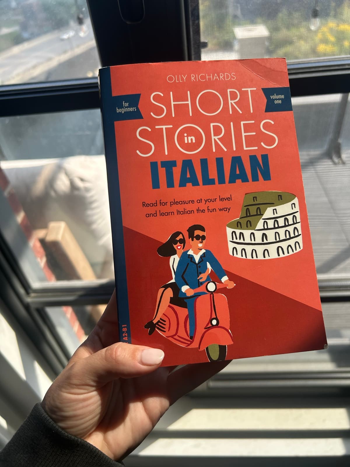A hand holding up a copy of Short Stories in Italian by Olly Richards, volume one, for beginners, in front of a sunny window